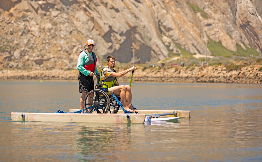 Member of the Cal Poly Adapted Paddling Program assisting a participant using a wheelchair adapted stand-up paddleboard while kayaking on calm water near a rocky shoreline.er near a rocky shoreline.