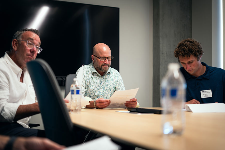 Three men seated around a conference table reviewing papers, with Dean Wendt reading a document in the center and McClain Kressman on the right.