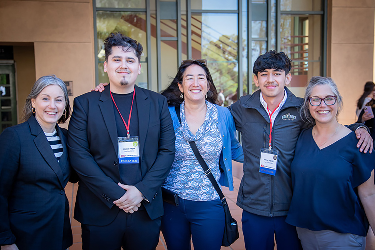 Group of biological sciences students and faculty mentor Professor Ale Yep posing together outdoors on campus in front of trees and landscaping.