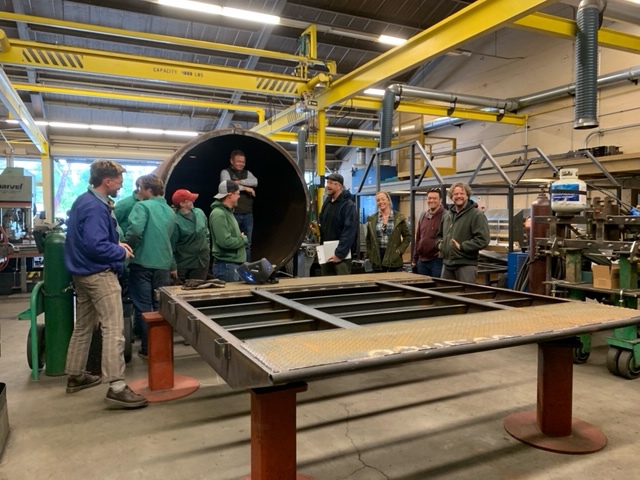 A group of people tour a metal fabrication shop, standing around a large steel cylinder and a worktable with a metal frame while industrial equipment and overhead cranes fill the workshop.
