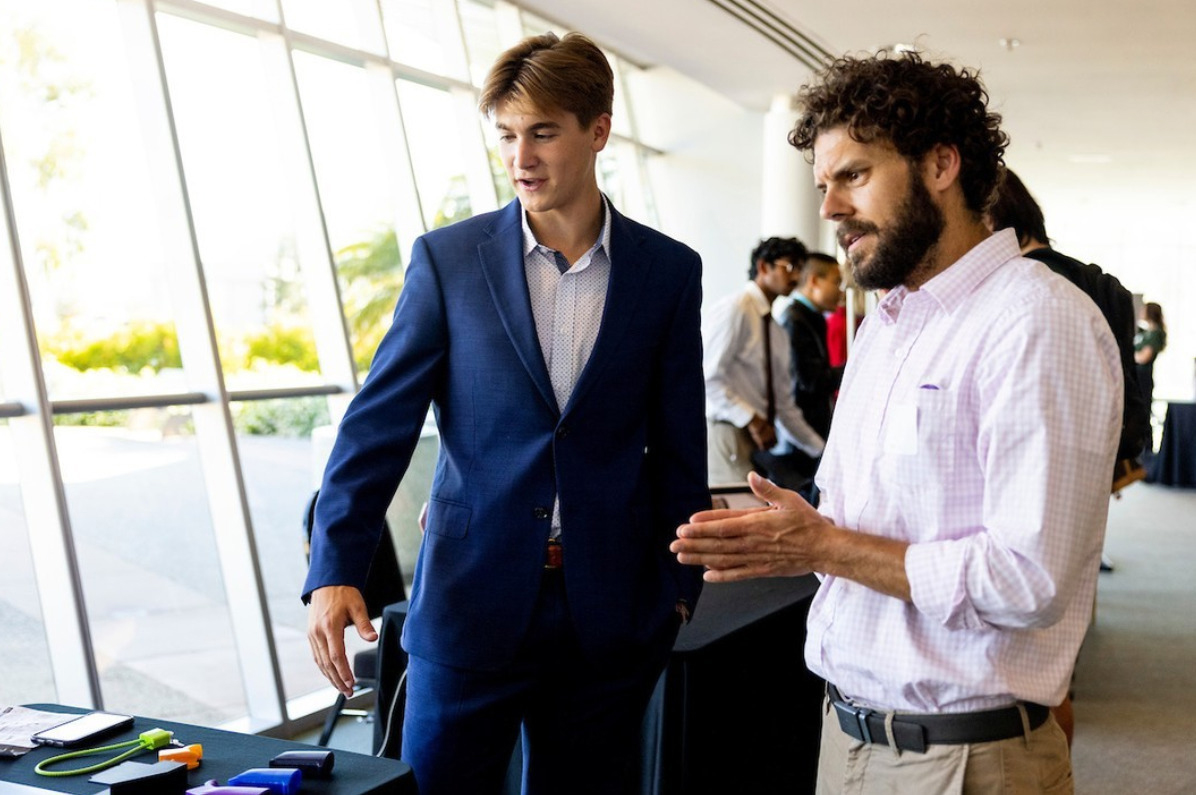 A student discusses a project with the faculty coordinator while presenting materials on a table during a research showcase.
