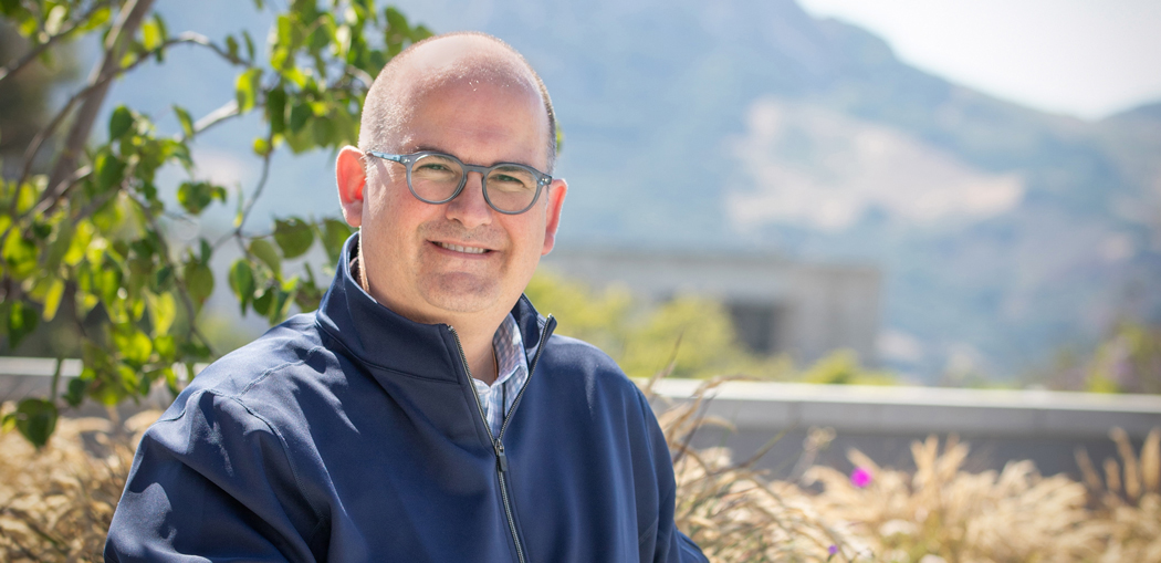 Daniel Kostalnick sits on a bench on the Cal Poly campus with trees and campus buildings in the background.