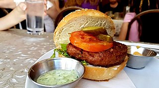 A plant-based burger with tomato and pickles is served on a bun with a side of sauce.