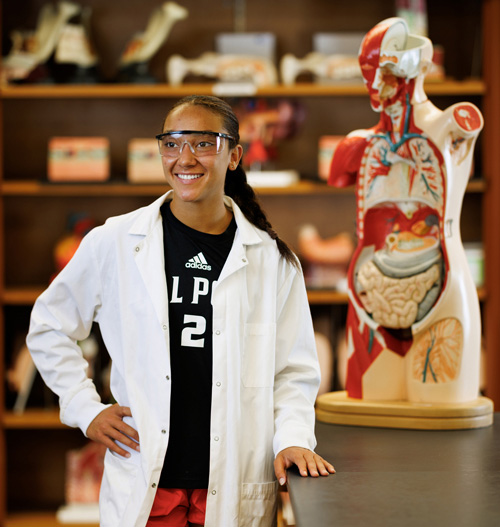 Cal Poly student Emma Brown, wearing safety glasses and a lab coat, stands beside a human anatomy model in a laboratory classroom.