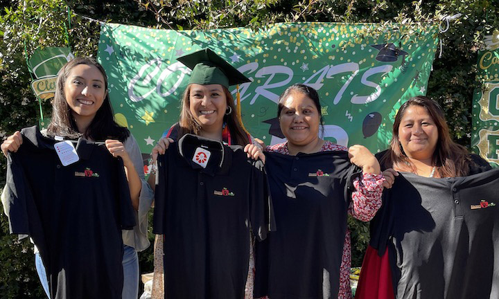 Four students, one wearing a graduation cap and gown, holding up black polo shirts in front of a Congrats sign.
