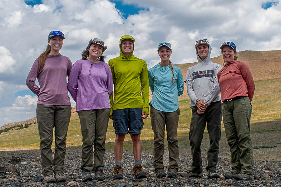 Group of researchers, including Brooke Wallasch (fourth from left), a Cal Poly master’s student in biological sciences, and Professor Dena Grossenbacher (far right) standing outdoors in a rocky alpine landscape with mountains visible in the background.