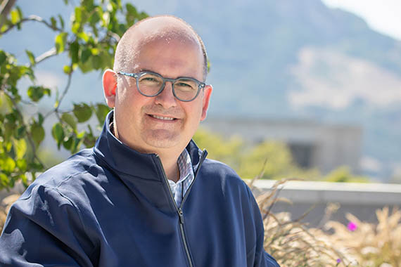 Daniel Kostalnick sits on a bench on the Cal Poly campus with trees and campus buildings in the background.