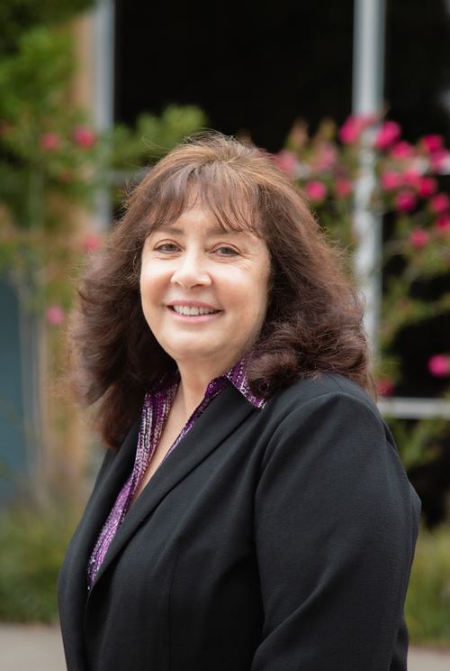 Sherri Brown smiles while standing outdoors with trees and greenery in the background.