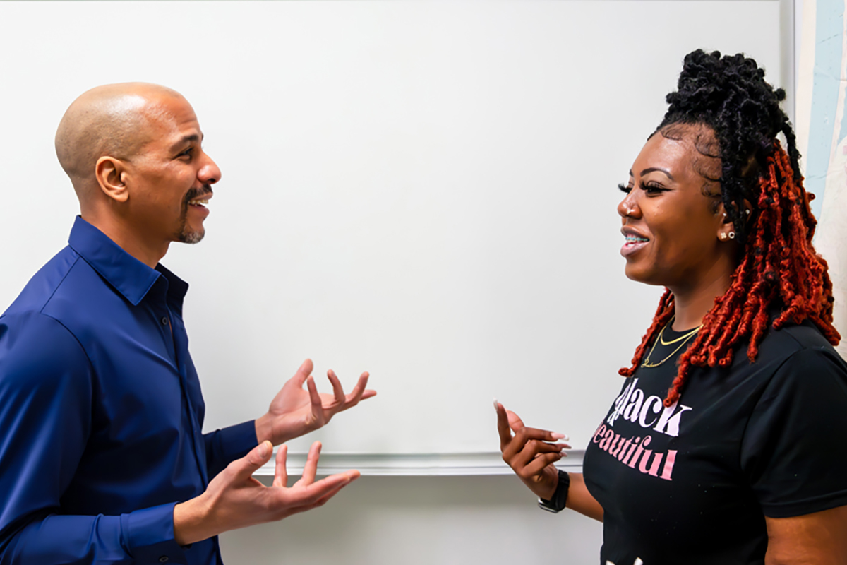 Cal Poly education professor Daniel J. Almeida and graduate student Shay Allen stand facing each other and talking in front of a classroom whiteboard.