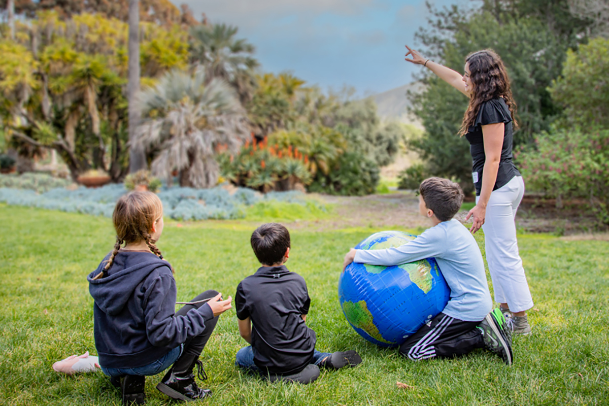 Instructor pointing toward plants while three children sit on the grass with a globe in a garden setting.