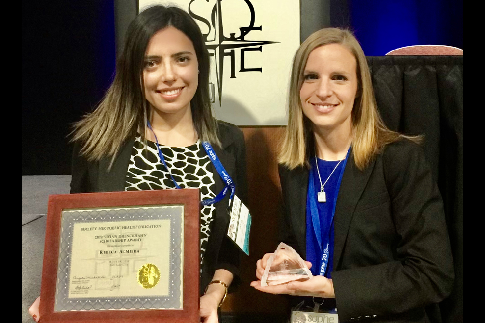 Rebeca Almeida and Julia Alber stand side by side at an event, smiling while holding an award plaque and a glass trophy.