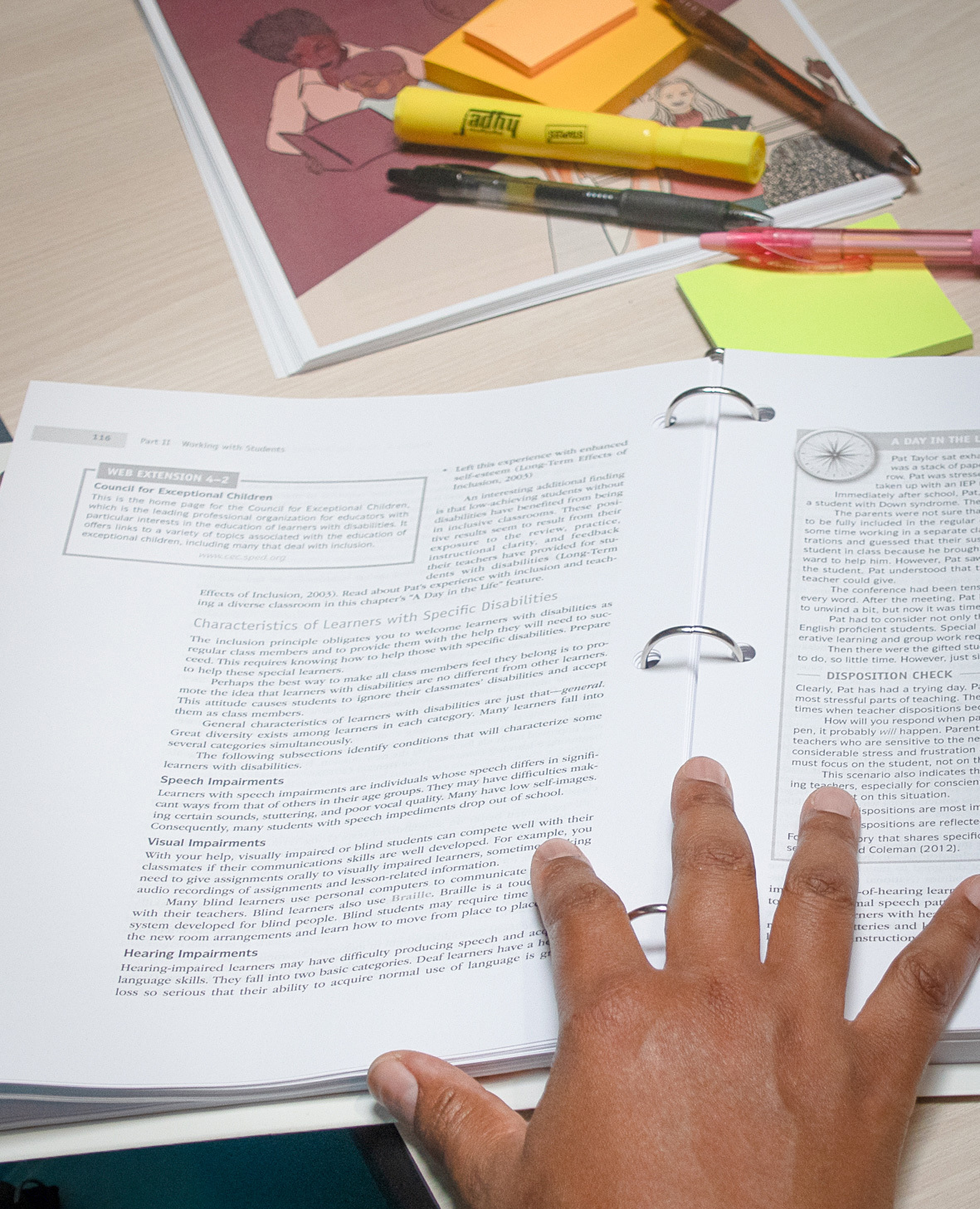 A hand rests on an open three-ring binder displaying textbook pages about learners with disabilities, with pens, sticky notes and a highlighter on the desk nearby.