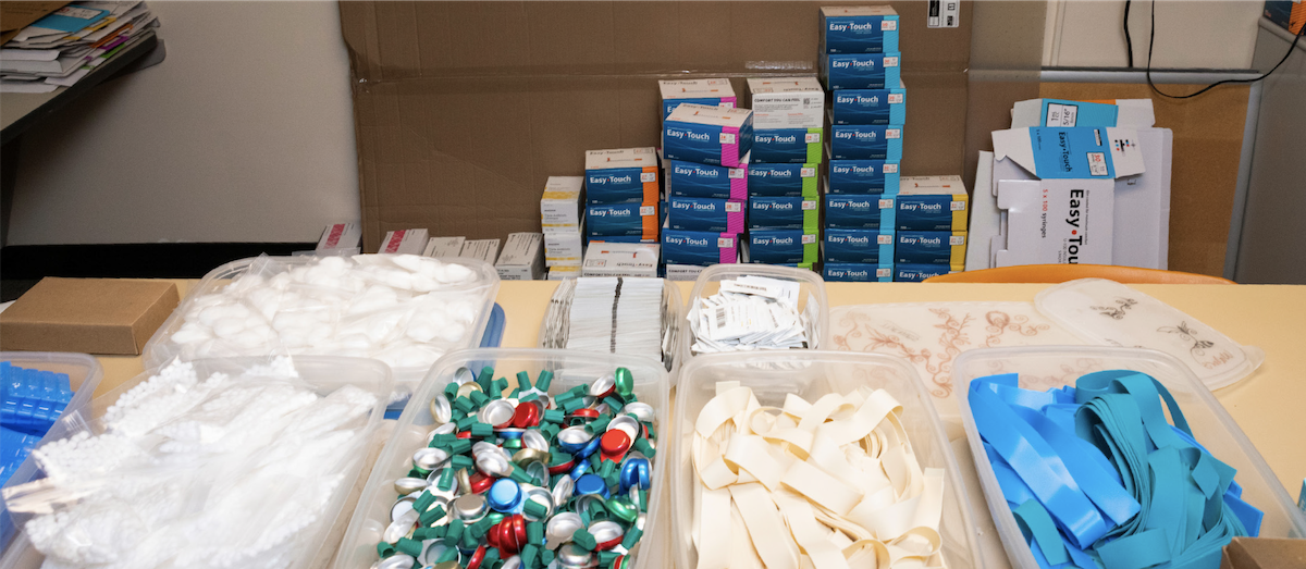 Hands place naloxone nasal spray doses beside a red overdose rescue kit on a table, with packaged harm-reduction supplies visible.