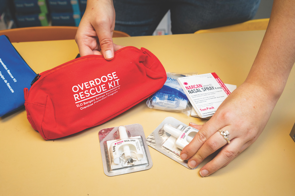 Hands place naloxone nasal spray doses beside a red overdose rescue kit on a table, with packaged harm-reduction supplies visible.