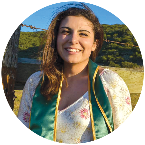 Micky Welther wearing a green graduation stole stands by a wooden fence with green hills behind her.