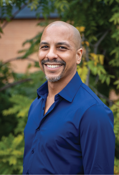 Portrait of Cal Poly education professor Daniel J. Almeida standing outdoors and smiling, with trees and greenery in the background.