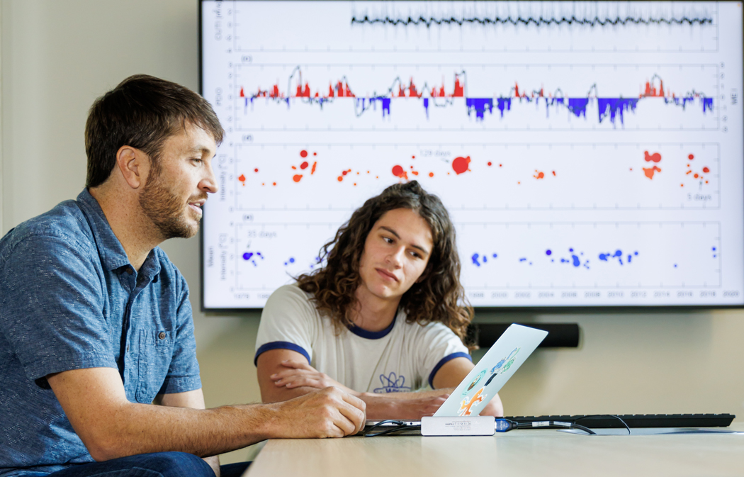 Ryan Walter and Michael Dalsin reviewing data on a laptop with graphs displayed on a screen behind them.