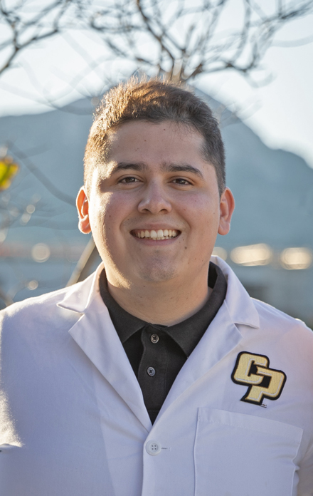 Portrait of Hector Reyes wearing a white lab coat with the Cal Poly logo, standing outdoors and smiling at the camera.