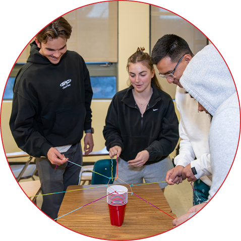 Students use colored strings and a rubber band to lift a stack of red plastic cups on a classroom table.