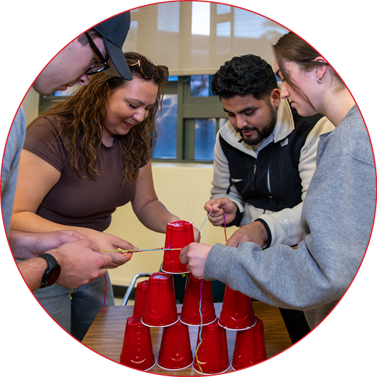 Four students lean over a classroom table, coordinating colored strings attached to a rubber band to add a red plastic cup to a pyramid of stacked cups