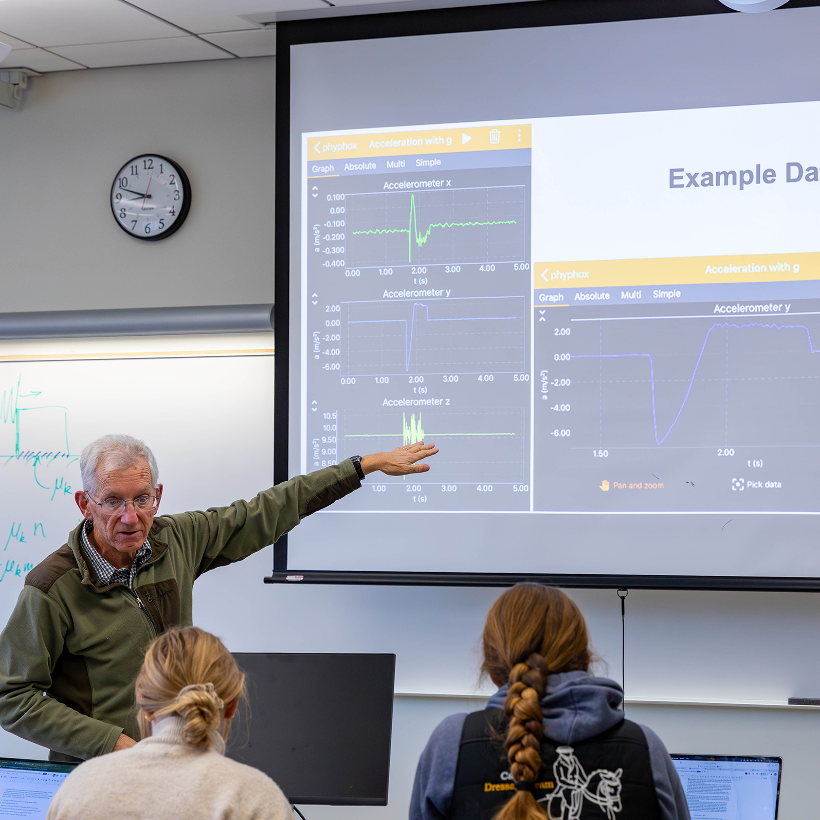 A professor stands beside a large screen displaying graphs and data, pointing to results while explaining the experiment to students.