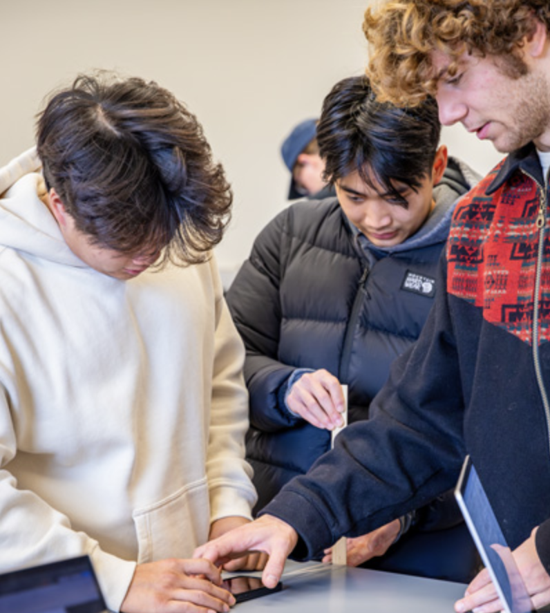Three Cal Poly students stand closely around a lab table, discussing results while using smartphones and equipment during a hands-on physics experiment.