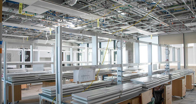 Bright laboratory inside the Frost Center with long workbenches, lab equipment and stools arranged beneath overhead utility lines and large windows.