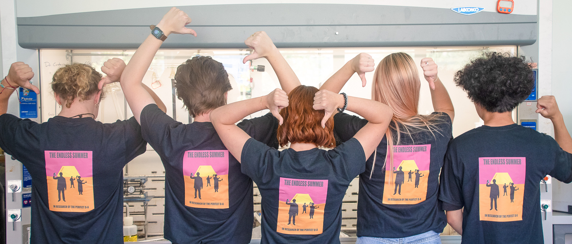 Six Cal Poly student researchers stand with their backs to the camera in a laboratory, pointing their thumbs toward matching lab T-shirts with a graphic on the back.