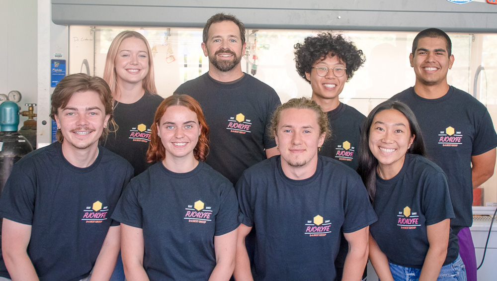 Cal Poly chemistry professor Phil Costanzo stands with student researchers from his lab group gathered together for a group photo in a laboratory setting.