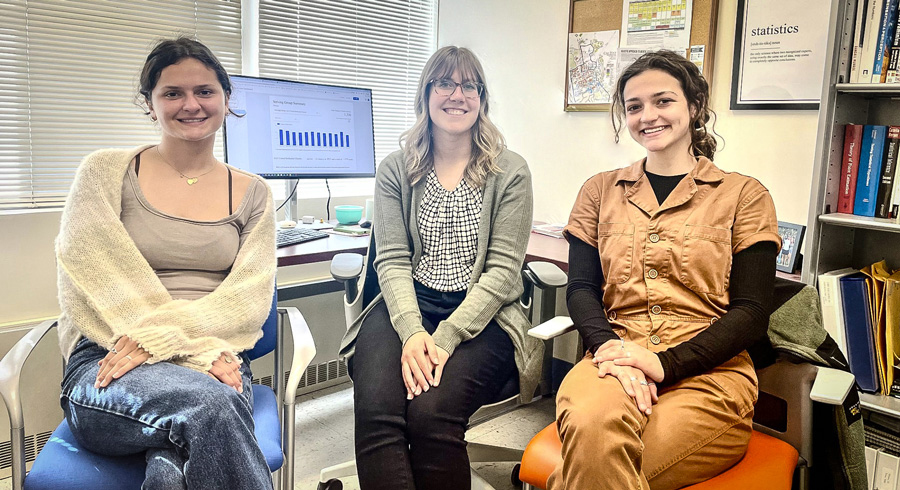 hree students sit side-by-side in an office, smiling toward the camera, with a computer monitor displaying a chart behind them and bookshelves nearby.