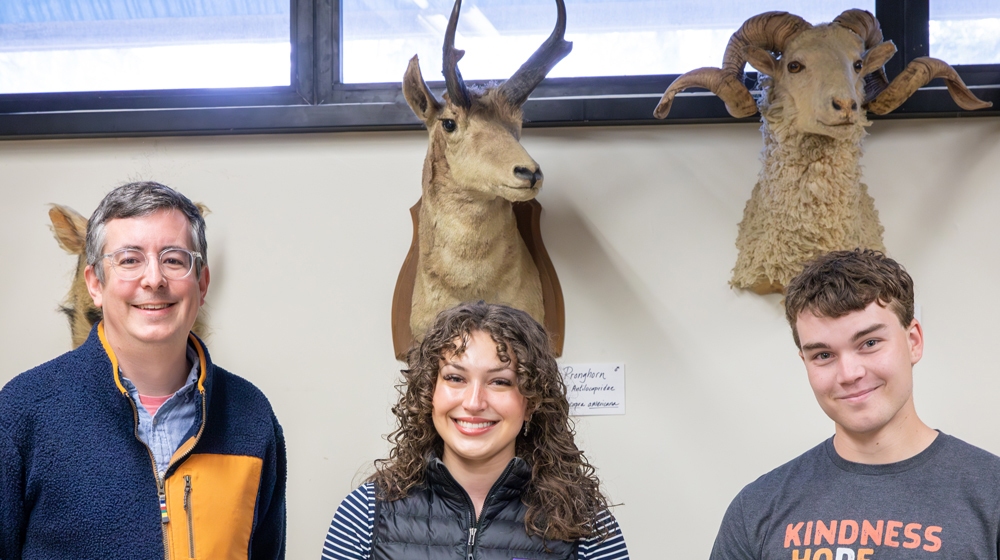 Three people stand indoors beneath mounted wildlife specimens, including pronghorn and bighorn sheep heads displayed on the wall behind them.