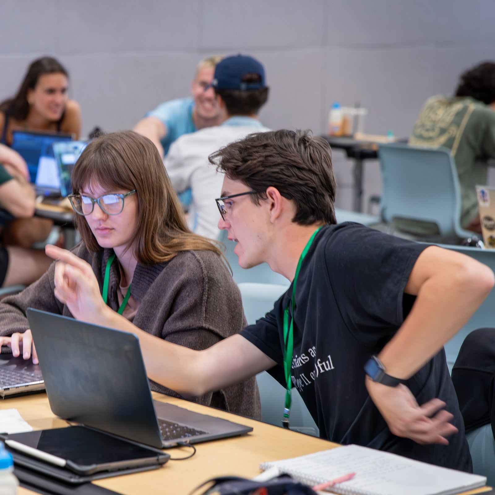 Two students sit next to each other in a crowded room with laptops in front of them. One student points to the other's screen, explaining something.