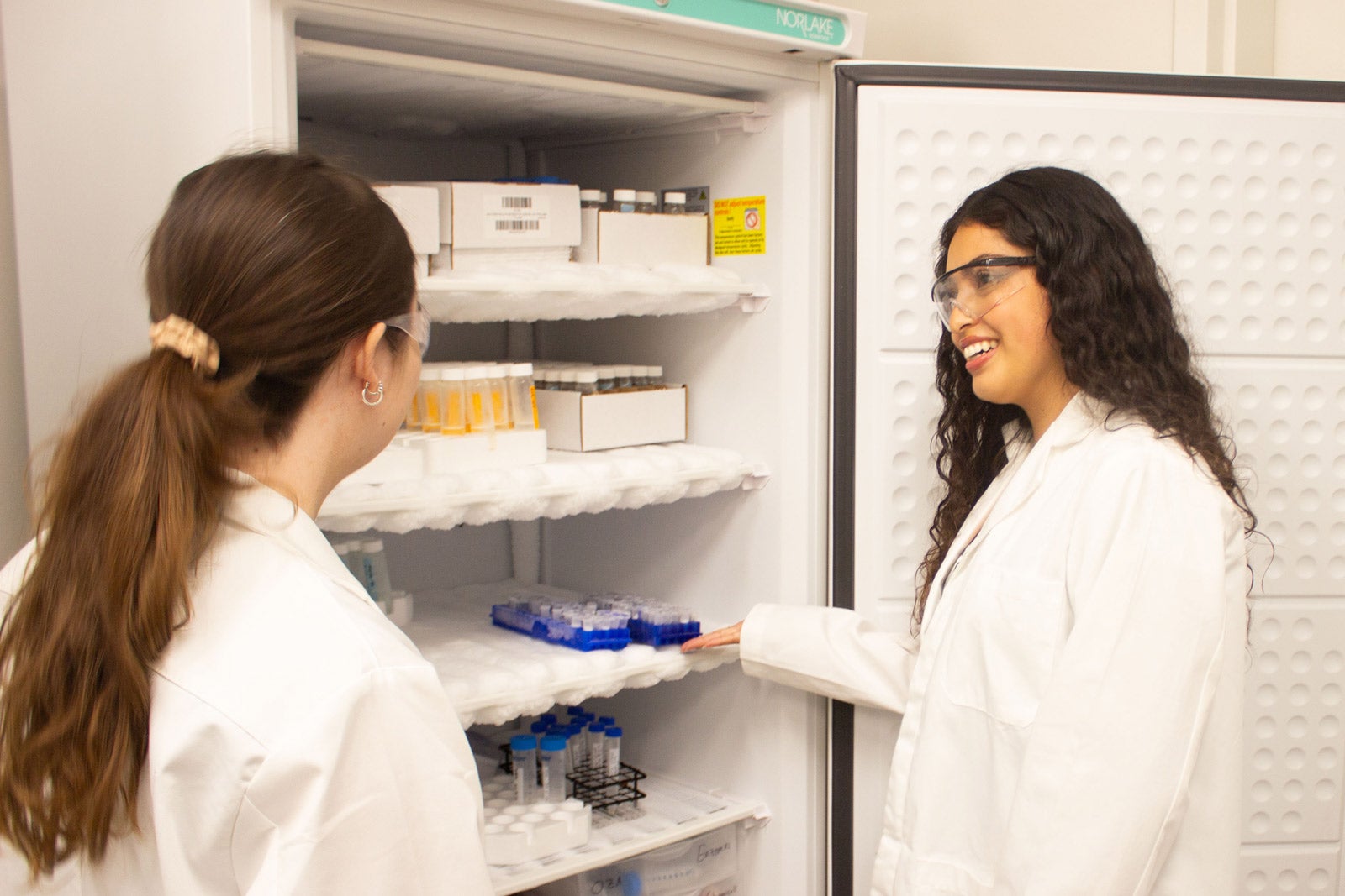 Two students wearing lab coats and safety goggles stand near frozen samples in a Frost Center lab.