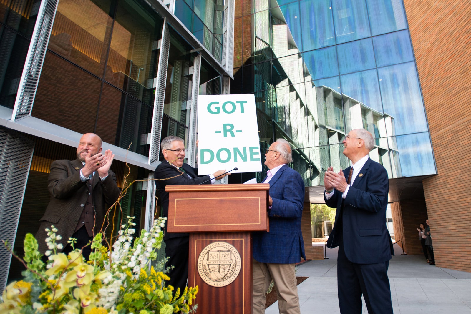 Cal Poly President Armstrong holds up a "Got-R-Done" sign at a podium during the William and Linda Frost Center for Research and Innovation Dedication Ceremony in 2023. Dean Wendt claps to the left, and Bill Frost and Phil Bailey stand to the right of the sign.