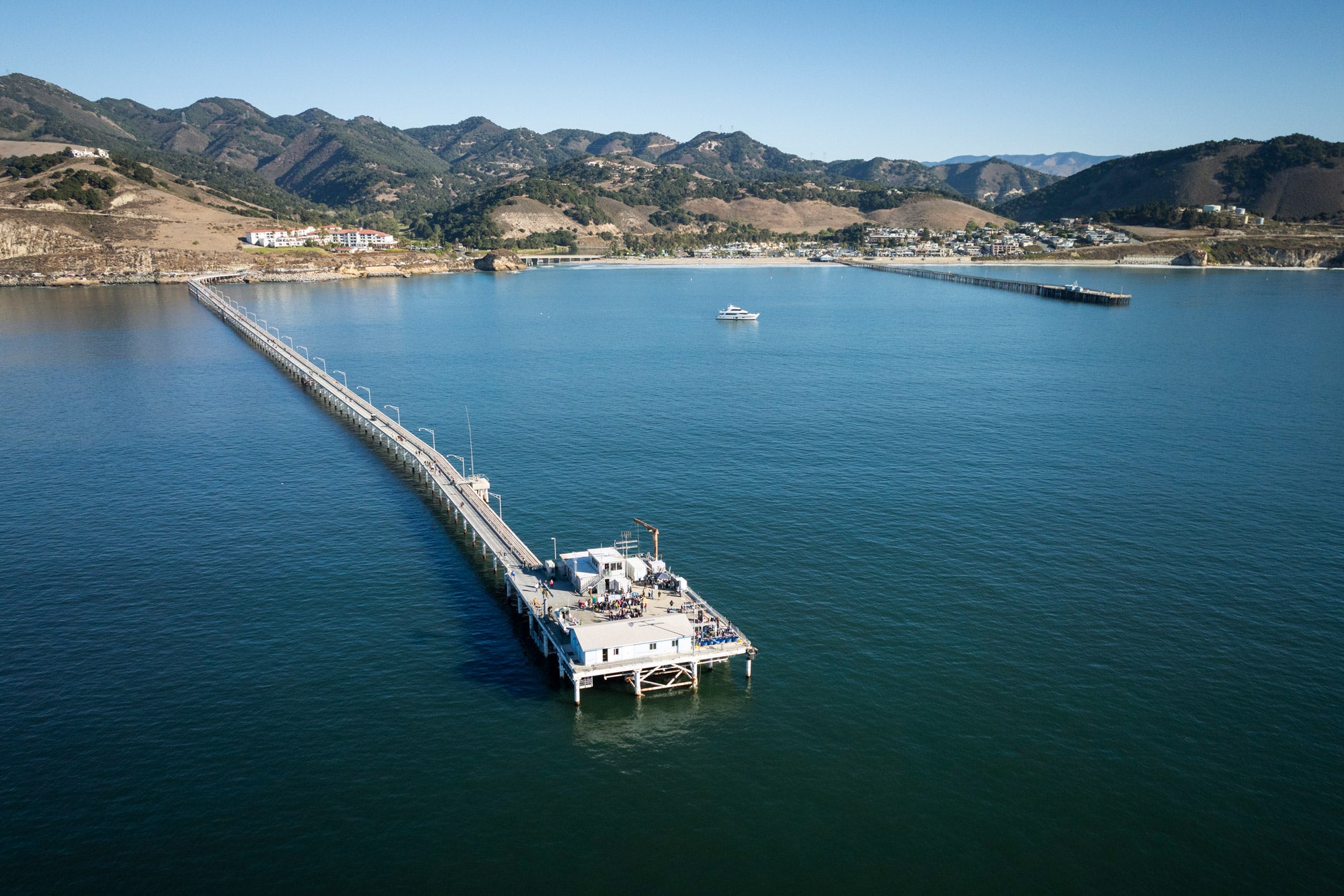 Wide angle drone shot of the Cal Poly Pier from above the water, looking back towards Avila.