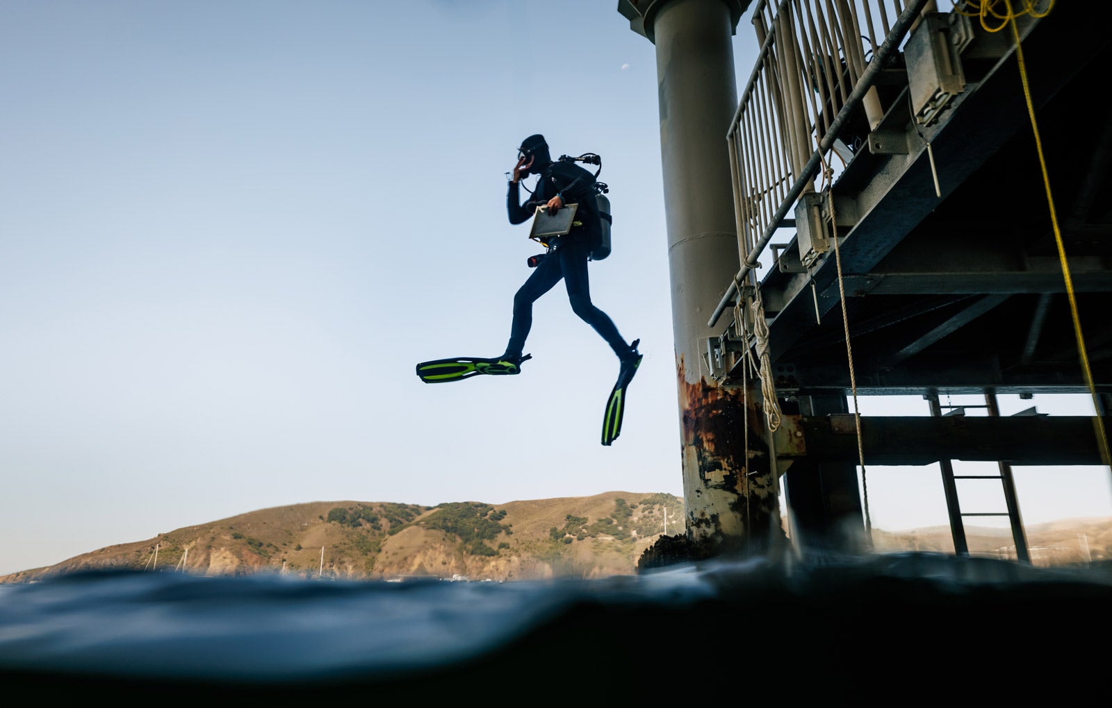 A student in scuba gear jumps off of the pier and into the ocean.
