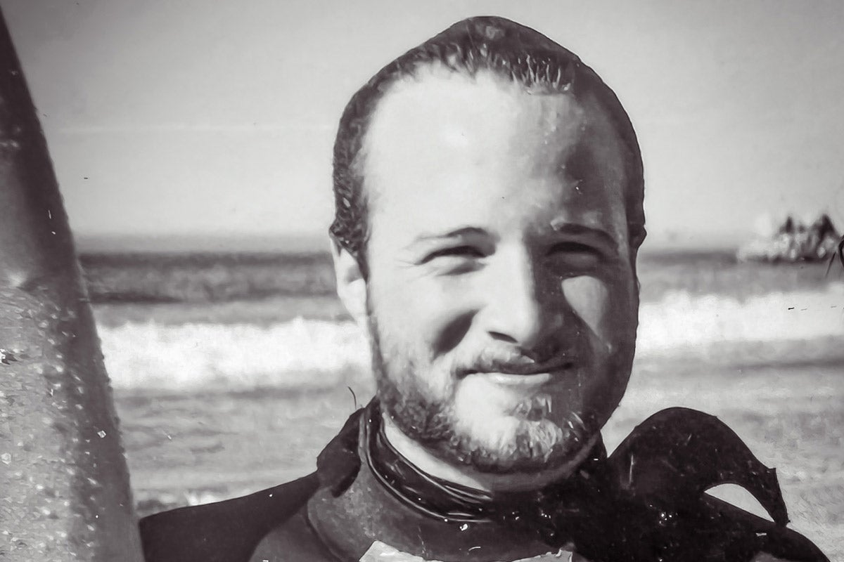 Black and white photo of Dean Wendt in 1990 as a marine biology student, wearing a wetsuit and holding a surfboard.