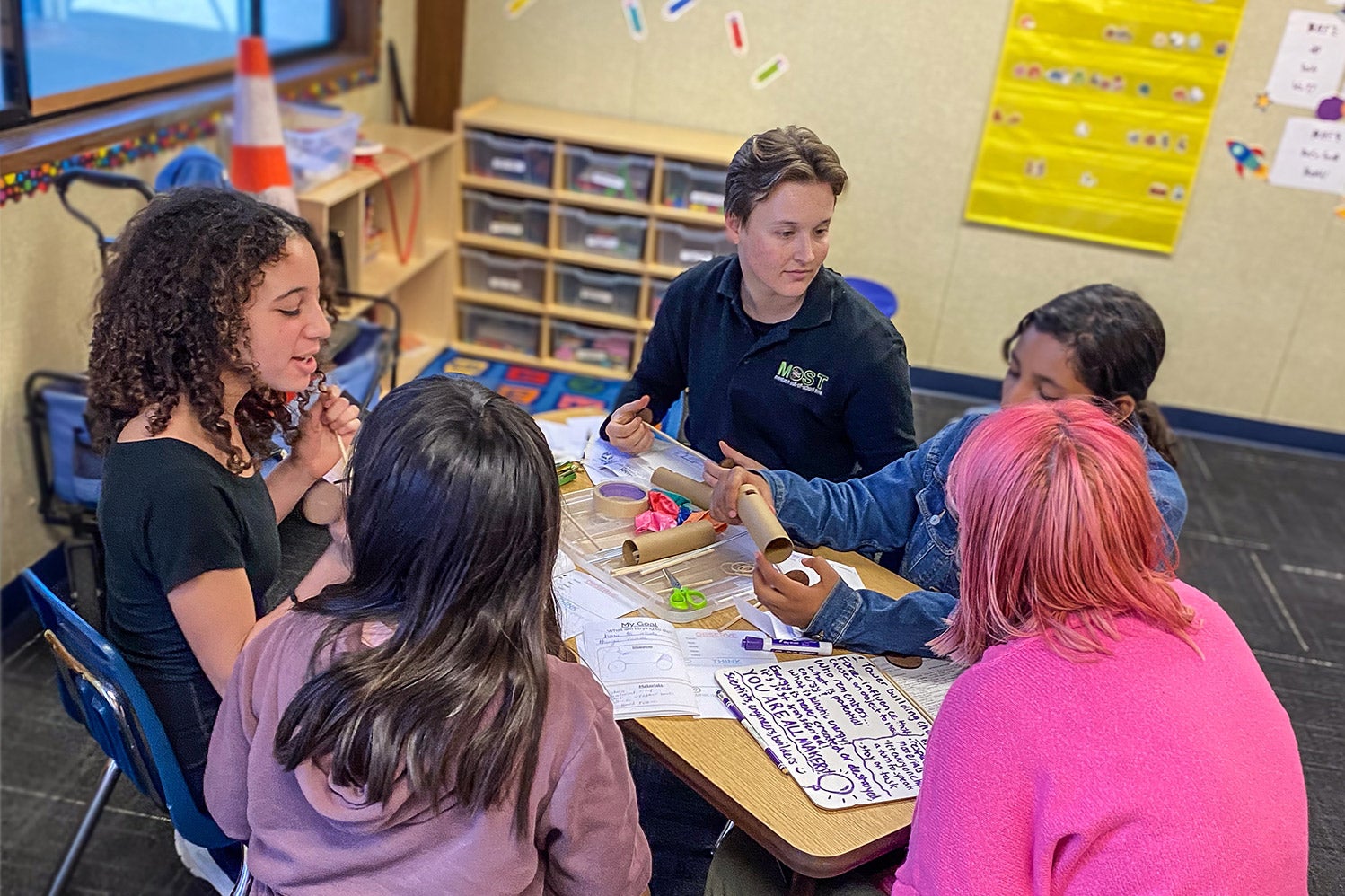 Claire Gillaspie, a liberal studies alumnus, speaks to elementary school students sitting at a classroom table, working together on a building activity.