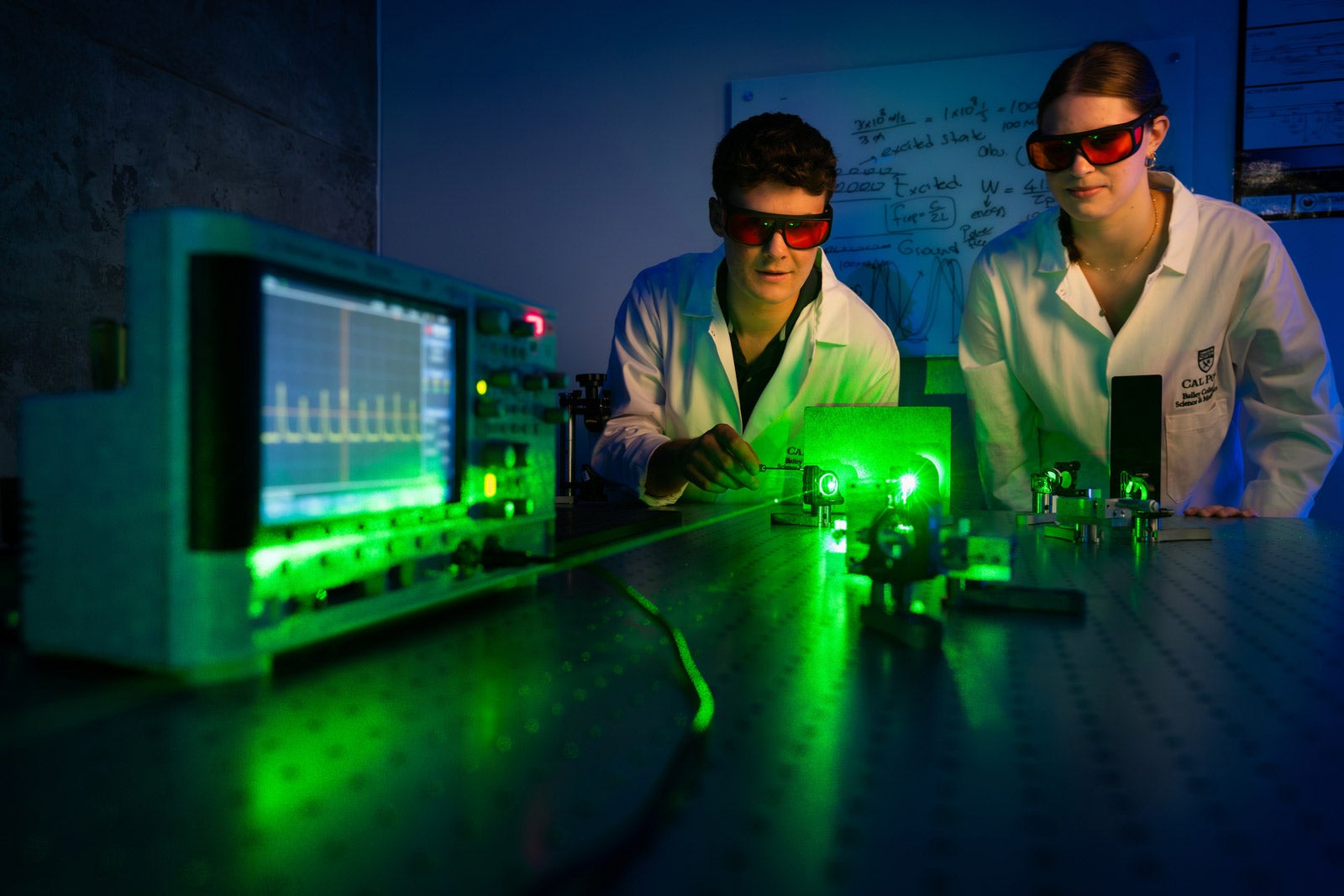 Students in lab coats and safety goggles examine the first femtosecond solid-state laser built at Cal Poly, which shines with a green light on the table.