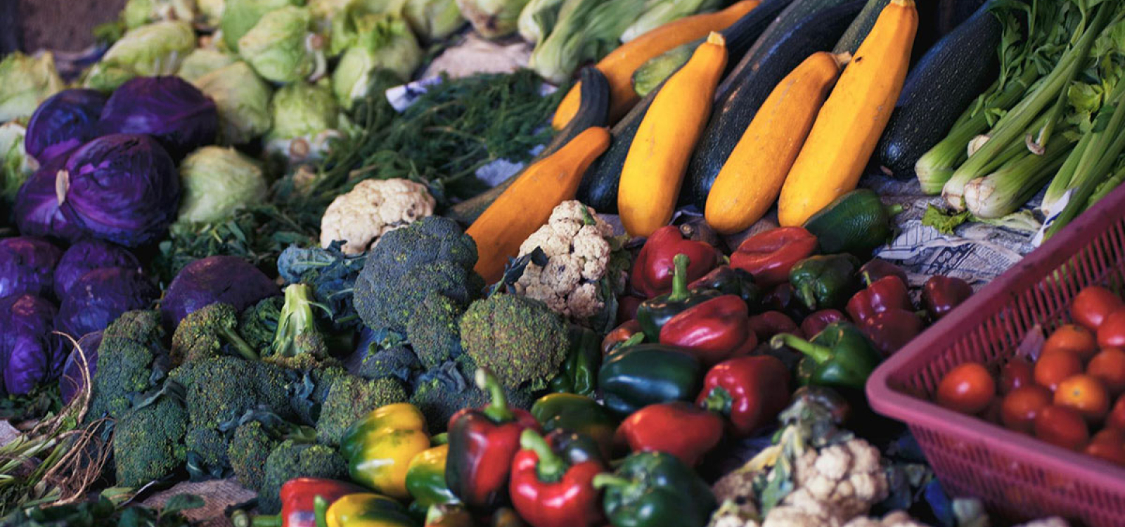 Display of fresh vegetables at a market stall, including zucchini, yellow squash, peppers, broccoli, cauliflower, cabbage and tomatoes.