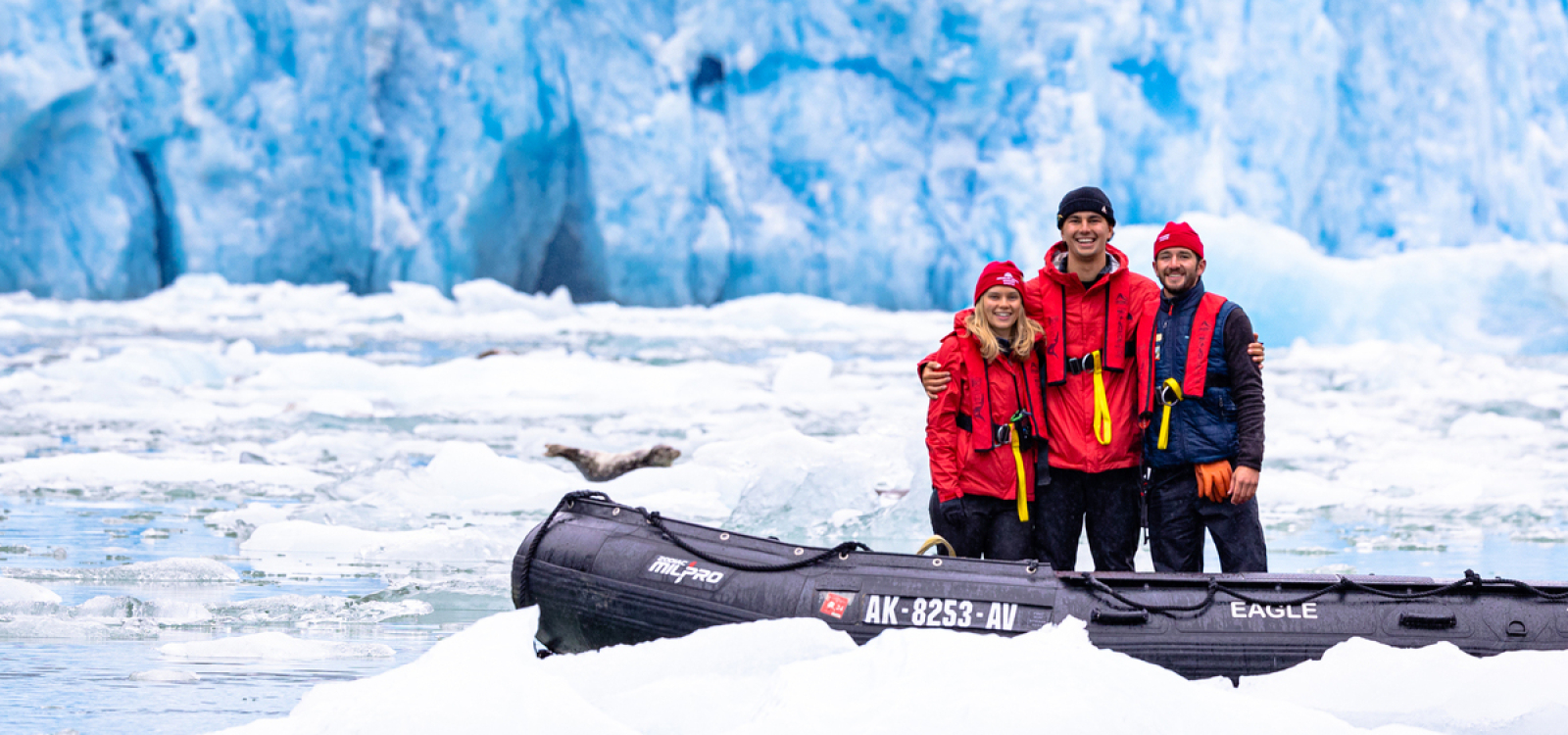 Cal Poly graduates Emily Thayer, Nathan Lesser, and Olin Bruton pose beside an inflatable boat on icy water with a blue glacier in the background during polar fieldwork.