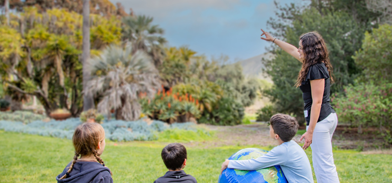 Instructor pointing toward plants while three children sit on the grass with a globe in a garden setting.