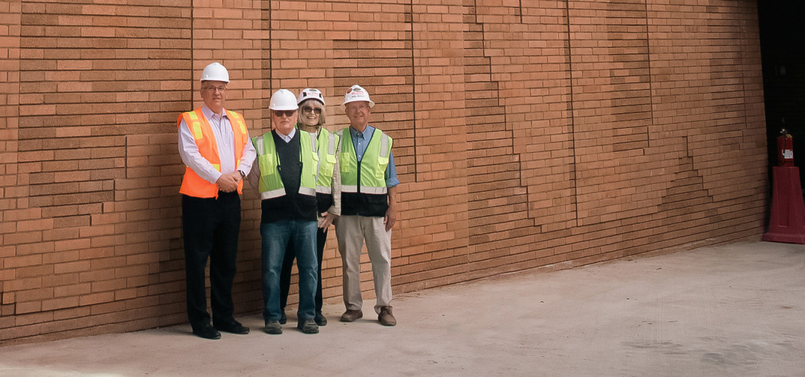 Cal Poly President Jeffrey D. Armstrong, Bill and Linda Frost, and Phil Bailey stand outside the Frost Center beside a tall brick wall spelling “Discere Faciendo.”