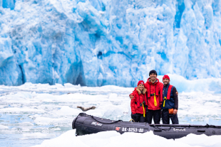 Cal Poly graduates Emily Thayer, Nathan Lesser, and Olin Bruton pose beside an inflatable boat on icy water with a blue glacier in the background during polar fieldwork.