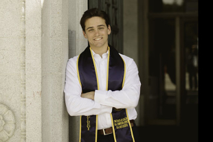 Nico Galin wearing a graduation stole and white shirt, leaning against a stone column outside a campus building.
