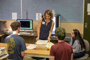 Katie Pearson picks up a piece of paper with a dried plant on it from a stack of more paper and plants. Three students watch her.