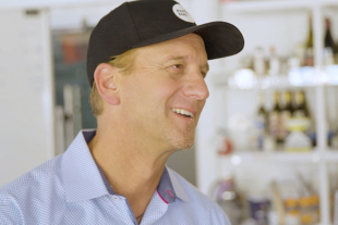 Tim Geistlinger (Biochemistry, ‘92), wearing a black cap and smiling while standing in a  retail space with bottles displayed on shelves behind him.