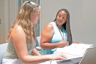 A Cal Poly professor and student sit at a table reviewing materials in a binder, discussing research during a meeting in a classroom.