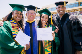 The Glover family with Dean Emeritus Phil Bailey stand together at a commencement ceremony, smiling and holding a diploma, with a dean in academic regalia; stadium seating and attendees visible in the background.
