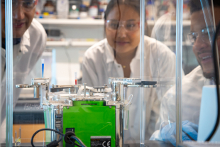 Three researchers in lab coats and safety glasses observe a laboratory instrument inside a protective enclosure, with small tubes of colored liquid samples positioned on the device.